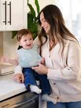Load image into Gallery viewer, Keep your baby’s clothes clean and dry during feeding time with a nontoxic, food-grade Silicone Bib from JuJuBe × Studio Oh! Made for babies 4 months and older, these bibs feature a deep front pocket to catch messes, and because they are dishwasher-safe, cleanup is fast and easy! Designed to fit comfortably and stay put, each bib features four ouch-free fasteners at the back of the neck strap, so it will stay comfortable as your baby grows. The Silicone Bib is BPA-free, PVC-free, phthalate-free, and lead-fr