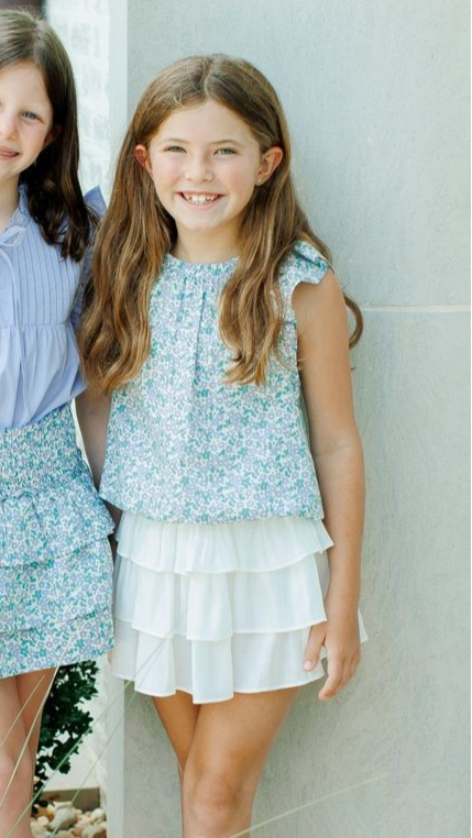 This girl's Allie Meadow Top from Pleat features a soft green and blue floral print, delicate ruffle shoulders, and cap sleeves for a charming and feminine look. The blousy feel adds comfort and ease, making it perfect for any occasion. Pair it with the coordinating tiered skirt for a complete and stylish outfit.