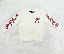 Load image into Gallery viewer, Elevate your little girl&#39;s wardrobe with our Girl&#39;s Red Bow Cardigan by Duffield Lane! This charming white knit cardigan features darling bows on both the sleeves and chest, adding a touch of sweetness to any outfit. Perfect for any occasion, this cardigan will keep her warm and stylish.
