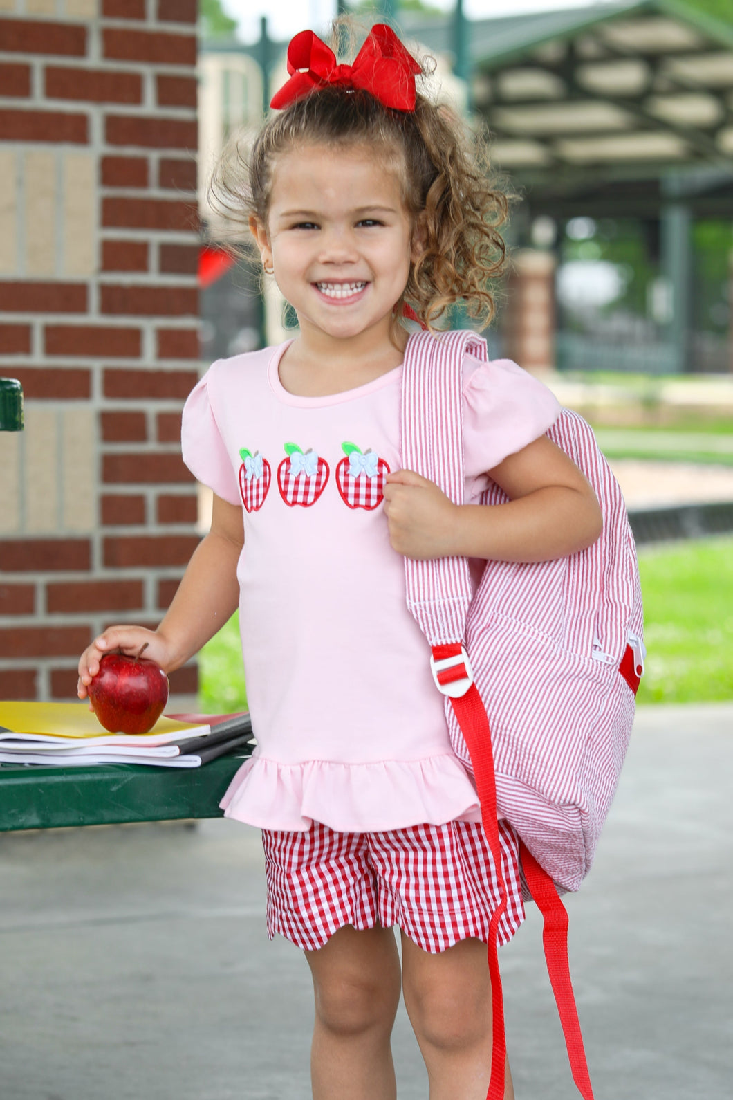 Delight your little one with this charming Toddler Girls Apple Trio Ruffle Short Set from Trotter Street. The pink ruffle hem tee showcases an adorable apple trio applique that celebrates the back-to-school season with style. Paired with eye-catching red and white checked shorts featuring a sweet scallop hem, this coordinated outfit offers both comfort and irresistible charm. Perfect for making a memorable impression on the first day of school or any summer playdate!