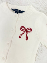 Load image into Gallery viewer, Elevate your little girl&#39;s wardrobe with our Girl&#39;s Red Bow Cardigan by Duffield Lane! This charming white knit cardigan features darling bows on both the sleeves and chest, adding a touch of sweetness to any outfit. Perfect for any occasion, this cardigan will keep her warm and stylish.

