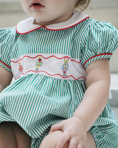 The Girls Nutcracker Kelly Bubble from The Yellow Lamb combines festive charm with elegant details, featuring green and white stripes complemented by an embroidered sash adorned with Nutcracker motifs. Accented with refined piping along the Peter Pan collar and sleeves, this bubble is a perfect choice for Christmas pictures and magical Santa visits, ensuring your little one looks both classic and captivating this holiday season.