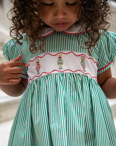 The Girls Nutcracker Kelly Dress from The Yellow Lamb combines festive charm with elegant details, featuring green and white stripes complemented by an embroidered sash adorned with Nutcracker motifs. Accented with refined piping along the Peter Pan collar and sleeves, this dress is a perfect choice for Christmas pictures and magical Santa visits, ensuring your little one looks both classic and captivating this holiday season.