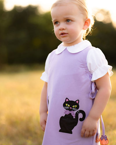The Emma Jumper Black Cats from The Yellow Lamb is a charming lavender shift dress perfect for your toddler’s Halloween celebrations. Featuring playful black cat appliques, this jumper combines festive fun with classic style, making it an ideal choice for little girls to enjoy the season in comfort and cuteness. Pairs with The Yellow Lamb matching Lavender Leggings so she can wear this as a dress or a set on cooler days!