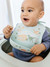 Load image into Gallery viewer, Keep your baby’s clothes clean and dry during feeding time with a nontoxic, food-grade Silicone Bib from JuJuBe × Studio Oh! Made for babies 4 months and older, these bibs feature a deep front pocket to catch messes, and because they are dishwasher-safe, cleanup is fast and easy! Designed to fit comfortably and stay put, each bib features four ouch-free fasteners at the back of the neck strap, so it will stay comfortable as your baby grows.
