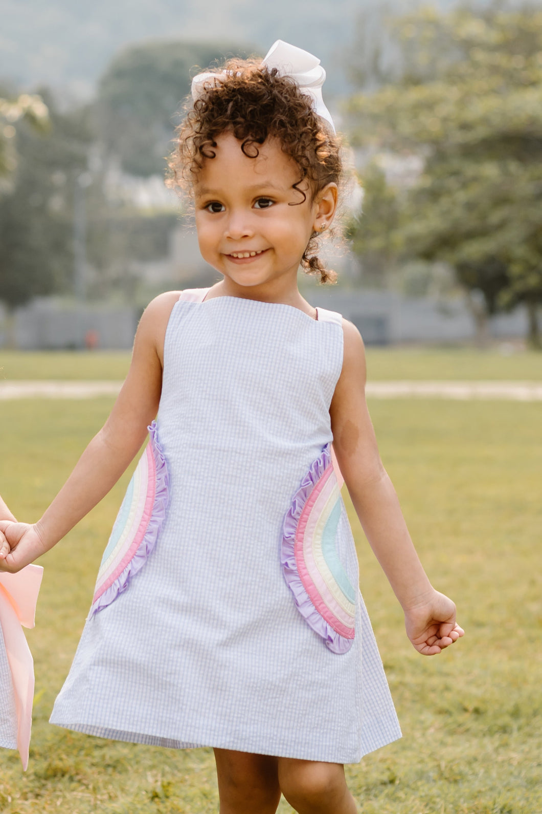 This adorable Rainbow Skye Dress from Zuccini Kids for toddler girls features mini light blue seersucker, vibrant rainbow appliqués on the pockets, and a colorful cross back with bow design. Perfect for any occasion, this dress will have your little one looking cute and stylish while staying comfortable.