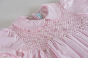 An utterly precious little girl's dress from Feltman Brothers! Scalloped stitching lines both the collar and puffed sleeves of this sweet looking dress, making it a perfect choice for your little darling. The smocking is covered in a beautiful diamond stitching and is embellished with hand embroidered clover flowers with a delicate pearl in the center of each. A zigzag design ends off the smocked bodice, and then continues into classy pleats down the length of the dress. Ties in the back with a lovely bow f