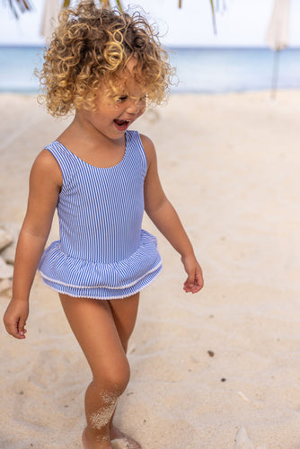 Bring the charm of Paris to the beach with this adorable skirted swimsuit. The blue and white seersucker stripes and white mini pompom accents give it a stylish and playful look. The airy open back with a bow detail adds a cute flair. Perfect for any fashionable toddler girl! From SnapperRock.